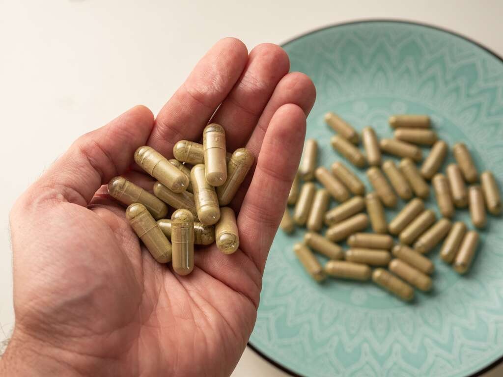 Hand holding kratom capsules with additional capsules on a teal plate